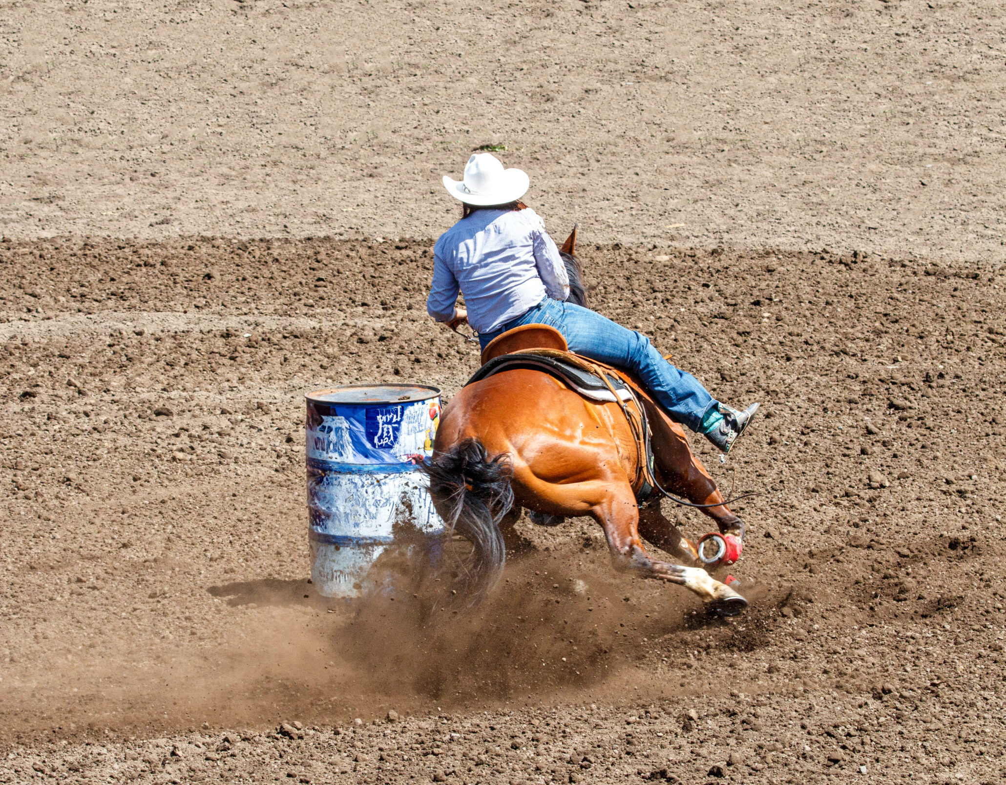 A cowgirl is riding a horse in a rodeo. She is competing in a barrel racing contest. The horse is kicking up a lot of dirt rounding the barrel. The horse is brown and the cowgirl is wearing blue.