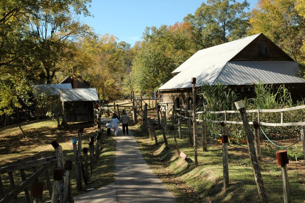 old-fashioned wooden buildings at end of a dirt road lined with rustic wooden fencing with a family walking down path