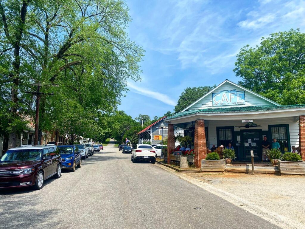 country road with trees on left, Whistle Stop Cafe on right, and cars parked down the lane