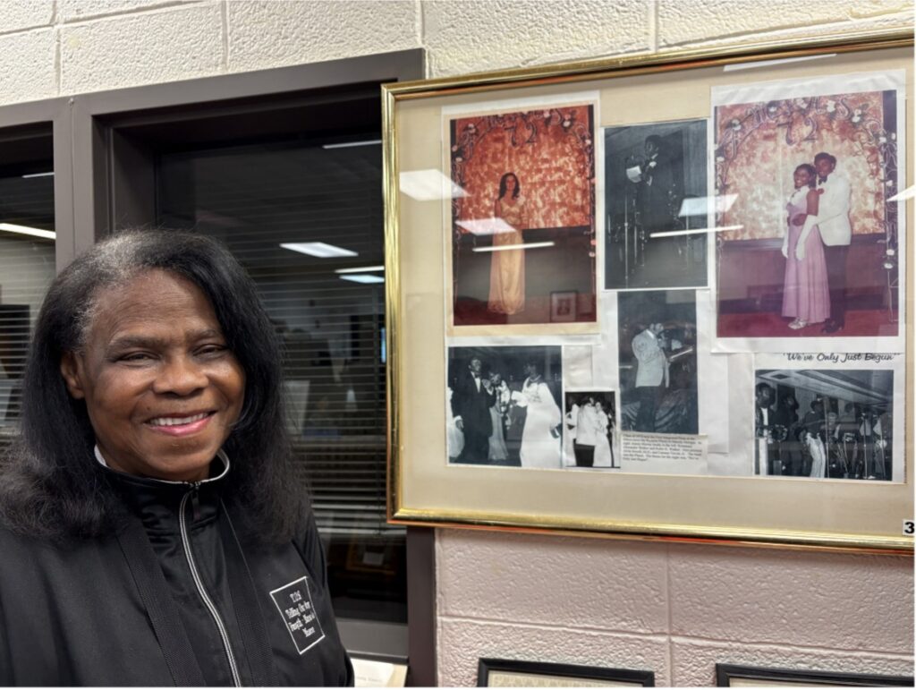 Older black woman with some gray in her shoulder length black hair stands beside a photo collage frame of images from her in high school