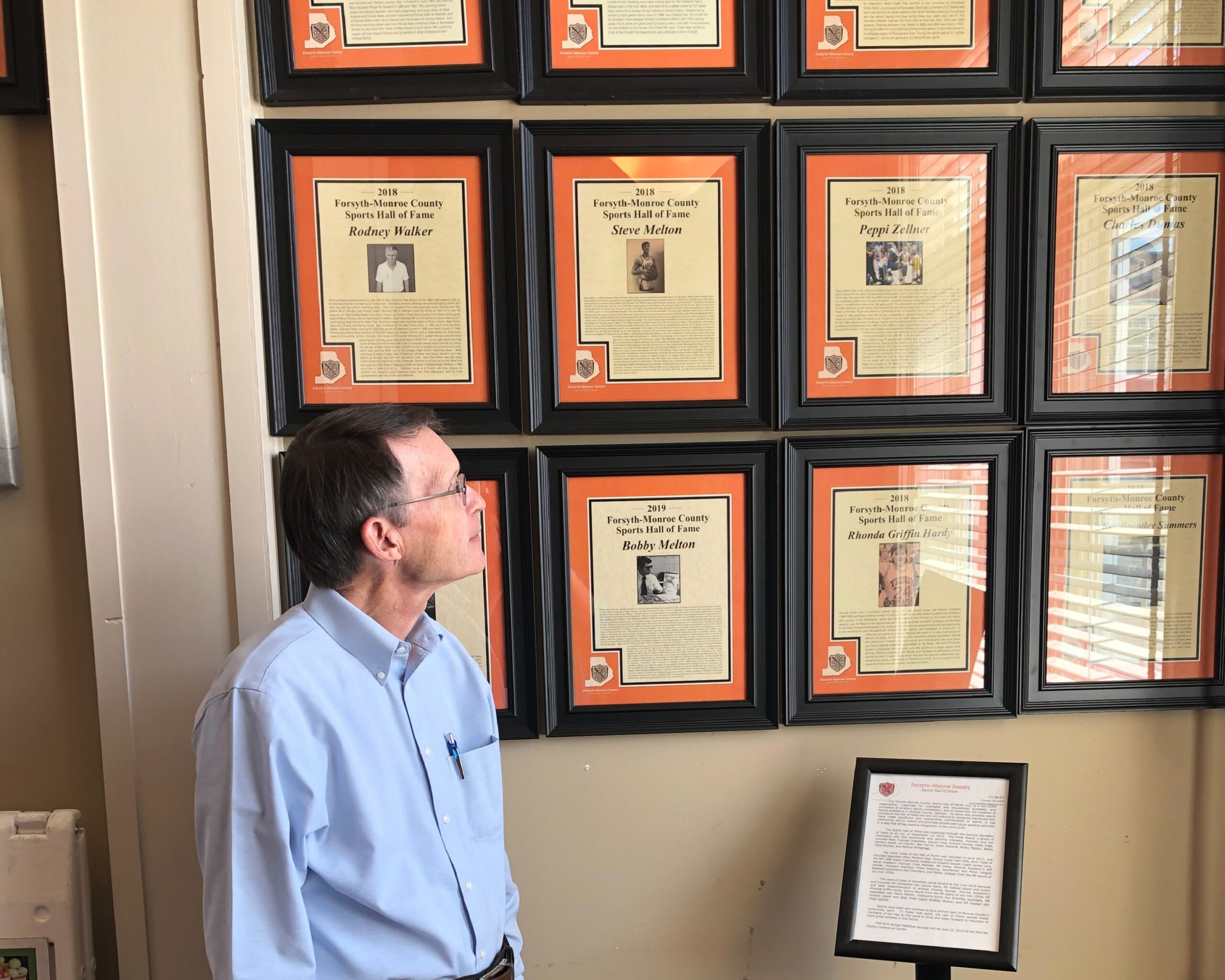 older man in glasses and light blue long sleeve collared shirt looks at a grid of plaques on a wall