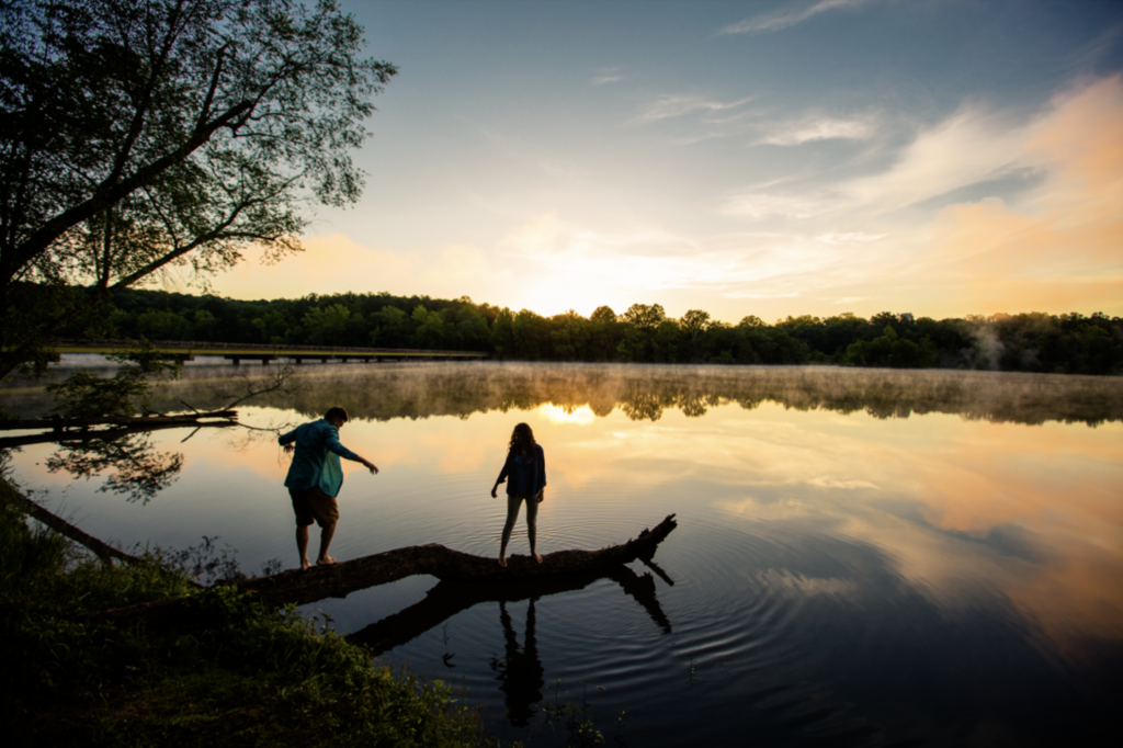 young adult man and woman balance on a tree that lays in a river at sunset