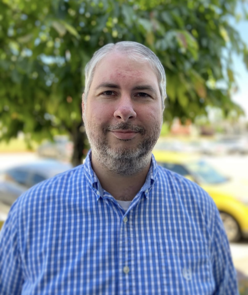 man with gray hair and short beard in blue collared shirt standing in front of a tree