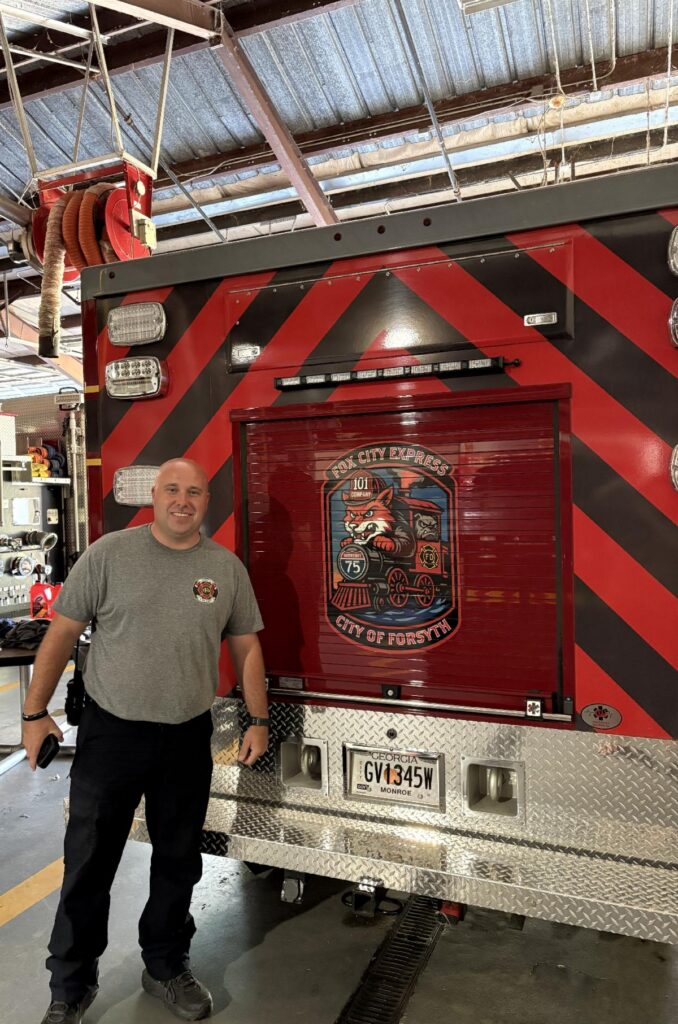 firefighter in tshirt stands in front of back of ambulance truck with badge design on back