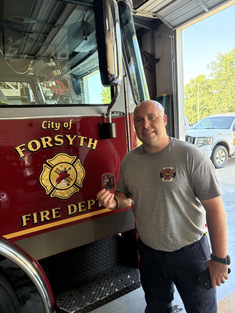firefighter in tshirt holds up sticker in front of fire truck