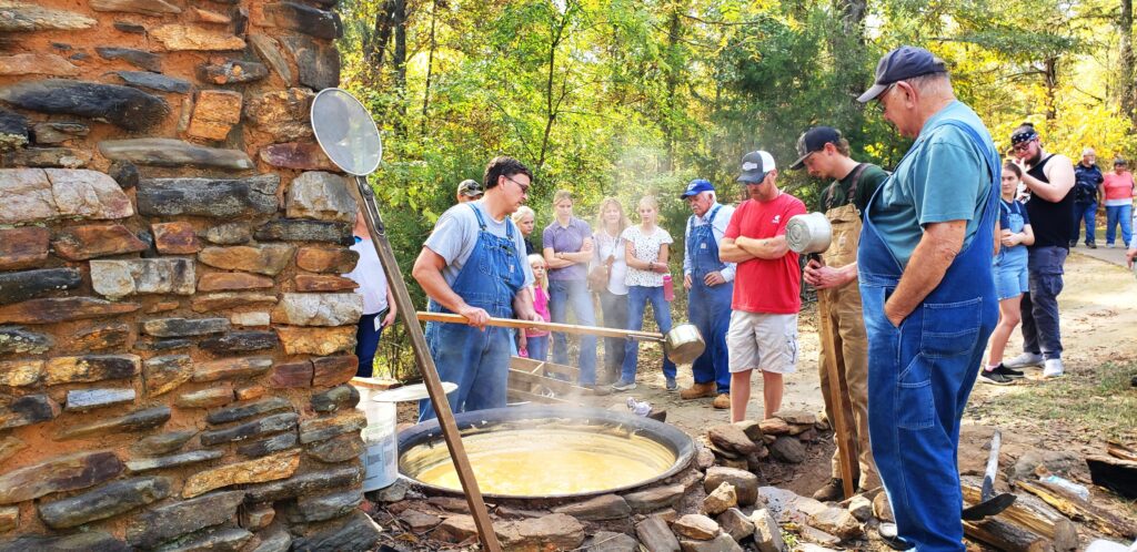 two men in overalls make syrup in historic outdoor pot surrounded by a crowd