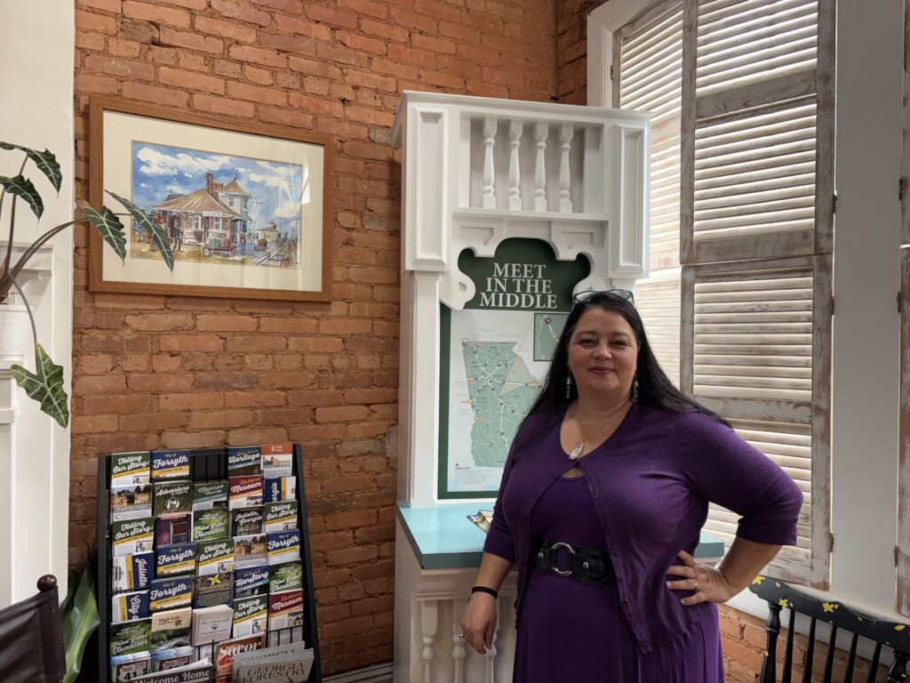 woman with long black hair wearing purple dress stands in front of an informational display and rack of brochures