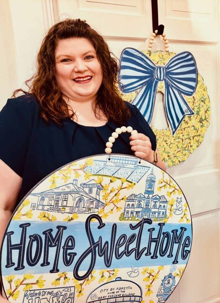 Nicole Newsom holding a “Home Sweet Home” sign decorated with Forsyth landmarks.